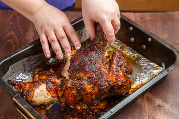 Women's hands cut off a leg from a whole baked chicken in a baking sheet.