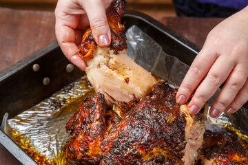 Women's hands tear off a ham from a whole baked chicken in a baking sheet.