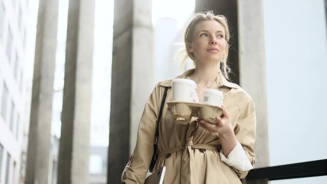 Portrait of pretty young blond businesswoman standing in office centre with takeaway coffee holder for her colleagues walking near modern business buildings outdoors Business morning concept