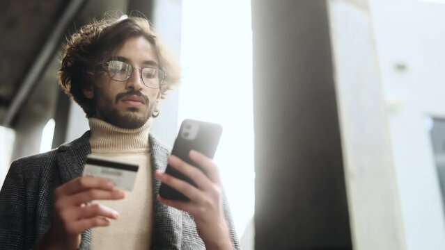 Portrait of confident arabian business man paying with credit card on smartphone in sunny modern buildings Successful young man doing payments online shopping in internet store easy pay outdoors