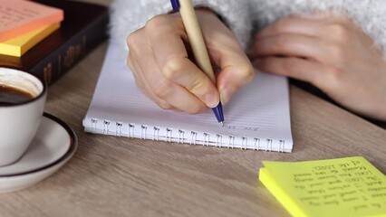 Human female hand writing on a notebook with pen on wooden table with closed bible book, a cup of coffee and colorful sticky notes. A closeup. Christian studying and reading holy Scriptures at home.