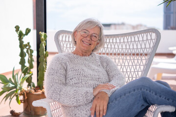 Portrait of senior attractive woman with cheerful smile relaxing at the armchair in the balcony at home looking at camera