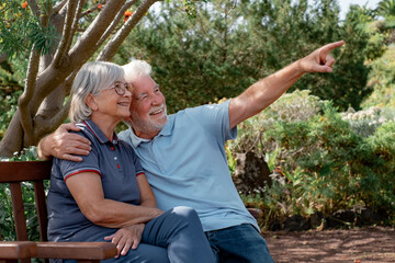 Happy senior couple in casual clothing sitting together on a bench enjoying sunny day in the garden