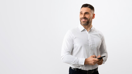 Great business app. Male entrepreneur with smartphone posing on grey background, looking aside at free space, panorama