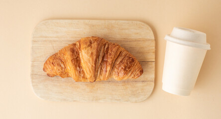 Top view of freshly baked croissant with golden crust next to a disposable paper cup of coffee. French traditional pastry on the beige background. Breakfast in the morning concept