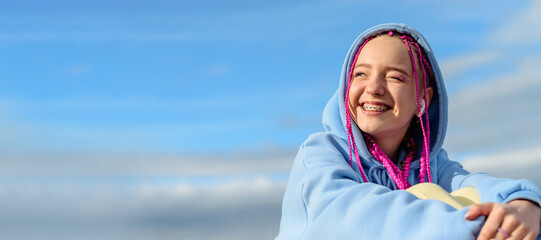 Portrait of a happy caucasian teenage girl with pink braids using wireless headphones against the blue sky.Technology,summer concept.Generation Z style.Copy space for text.