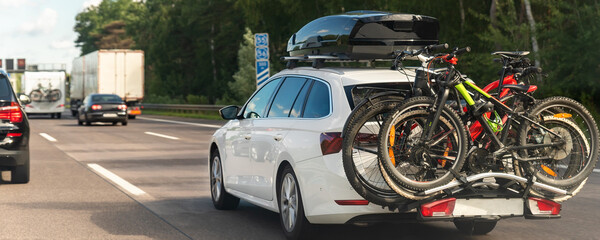 Back view modern white family wagon car with mounted roof box trunk and bike tail carrier driving european highway road against blue sky summer day. Lifestyle travel adventure trip journey concept