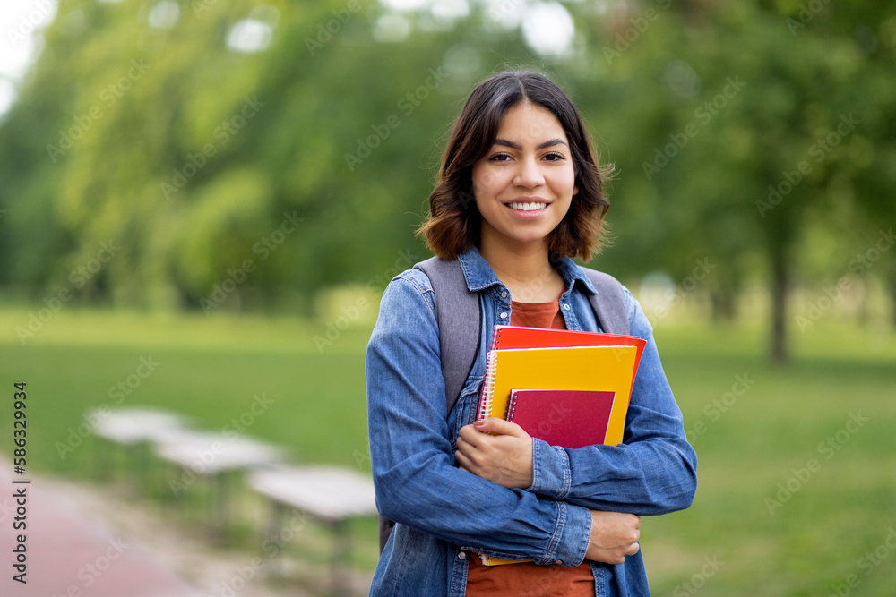 Wall mural smiling beautiful middle eastern female student holding workbooks while posing outdoors