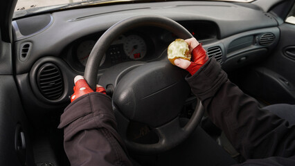  hands on the wheel of a car in red gloves, with an apple in one hand a person drives a car. Concept: Eating while driving a car   - obrazy, fototapety, plakaty