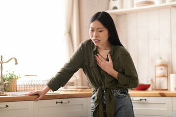 Sick chinese woman suffering from heartburn in kitchen, touching chest - obrazy, fototapety, plakaty