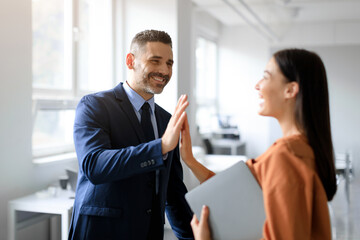 Fototapeta premium Happy coworkers celebrating achievement in office, man and woman giving high five and smiling, enjoying job well done