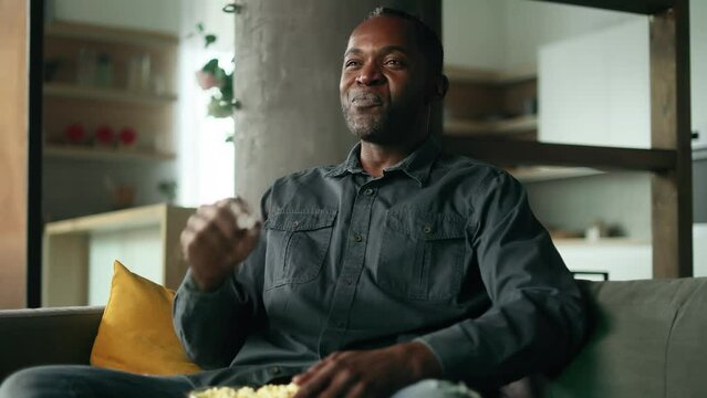 Portrait of happy positive mature african american man with popcorn laughing while watching comedy movie show serial enjoying leisure time at home alone Fun weekend time