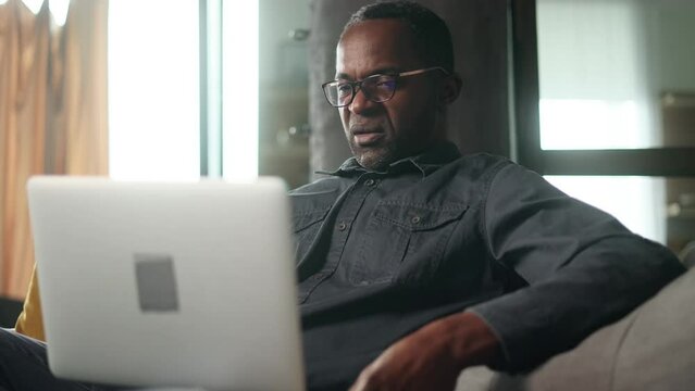 Boring mature african american man scrolling feed of social networks products in internet store on laptop in the living room Tired exhausted male look at computer screen without enthusiasm at home