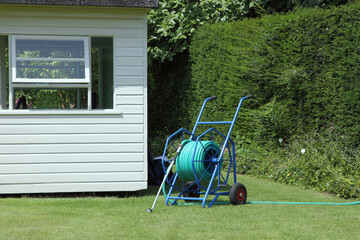 Garden hose pipe reel on wheels, ready for watering plants and lawn, next to a white, wooden shed . - 586335342
