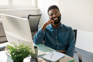 Portrait of happy black male ceo sitting at workplace and smiling at camera, working in office in front of computer