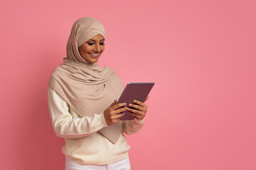 Fototapeta premium Smiling Muslim Woman In Hijab Using Digital Tablet, Standing Over Pink Background
