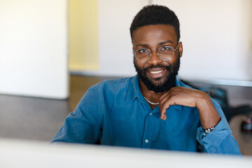 Portrait of happy black businessman sitting at desk with computer, working in office and smiling at camera, copy space