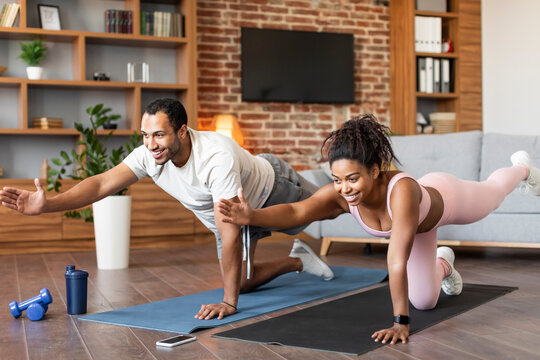 Sport lifestyle, family body care at home. Smiling young black guy and female in sportswear doing leg exercises