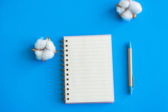 open notepad and craft pen next to it on a blue background. several cotton flowers nearby. place for text. view from above