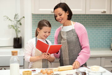 Glad caucasian young mom and small daughter in aprons make cookie dough, look at recipe book in...