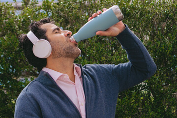 Middle-aged Latin man drinking water from a reusable thermos bottle while wearing headphones. - obrazy, fototapety, plakaty