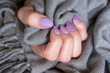 Close-up of well-groomed female nails with lilac color gel. Beautiful clear fingers of a young girl with a lilac-purple manicure. Brilliant colors and hands gripping a gray dress.
