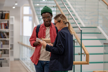 Two smiling diverse students talking discussing homework while standing at university library, caucasian girl and african american guy studying together, communicating in college campus
