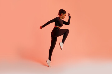 Fototapeta na wymiar Athletic black lady jumping in mid air, training over peach neon studio background, full length, side view, copy space