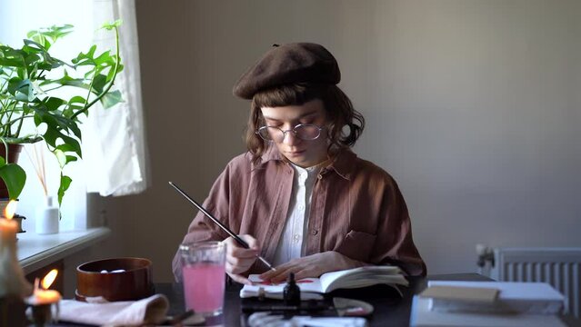 Focused millennial girl artist wearing beret and glasses sitting at table drawing at home. Concentrated creative young woman holding paint brush painting in studio. Art, creativity and inspiration
