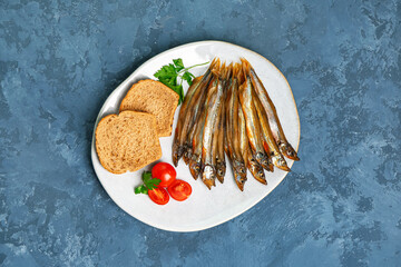 Plate with delicious smoked capelin, bread pieces and tomatoes on blue grunge background