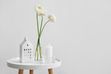 Burning candle, vase with flowers and house holder on table near light wall