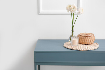 Burning candle, basket and vase with flowers on table near light wall