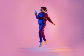 Full body length shot of young black lady in sportswear jumping and using cellphone over pink neon background - obrazy, fototapety, plakaty