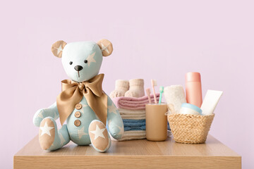 Toy bear with baby clothes and accessories on table near lilac wall