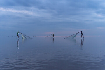 Fototapeta premium Silhouette of Vietnamese fisherman holding a net for catching freshwater fish in nature lake or river with reflection in morning time in Asia in Vietnam. People.