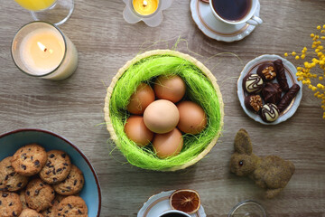 Easter eggs in the basket, bowl of cookies, chocolate pralines, Easter bunny figurine, cups of tea, glasses of juice, flowers and lit candles on the table. Top view. 