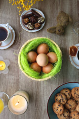 Easter eggs in the basket, bowl of cookies, chocolate pralines, Easter bunny figurine, cups of tea, glasses of juice, flowers and lit candles on the table. Top view. 