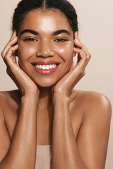 Beauty face. Smiling african american woman touching healthy skin portrait. Beautiful happy girl model with fresh glowing hydrated facial skin and natural makeup on brown background at studio