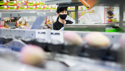Obraz premium Focused diligent efficient glad female worker wearing protective face mask working at fruit warehouse carrying box with mangos