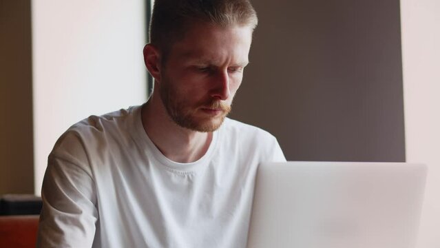 Freelancer sits in a cafe with a cup of coffee and works on a laptop. A frowning man looks at a laptop screen and types something.