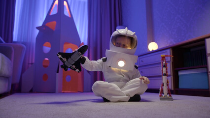 Small child in an astronaut spacesuit plays at home with toy Space Shuttle orbiter. Boy dreams of becoming an astronaut and flying into space. Dream is to fly in space