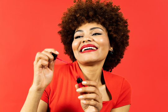 A dark-skinned girl with afro hair posing on a red background with makeup products, the woman looks sideways happily as she is wetting the liquid lipstick brush to touch up her lips. Makeup concept.