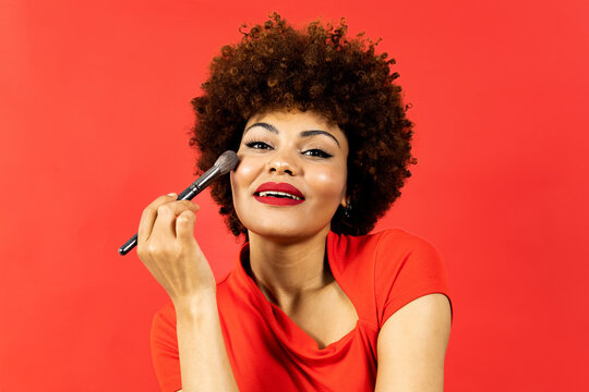 Portrait of a dark-skinned girl with afro hair posing on a red background with a powdery makeup brush approaching her face as she looks into the camera. Concept of makeup in African people