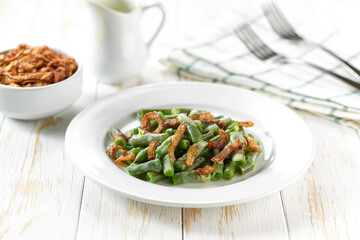 Baked green bean with crispy fried shallots and cream sauce in a ceramic plate on a white wooden table, close up.