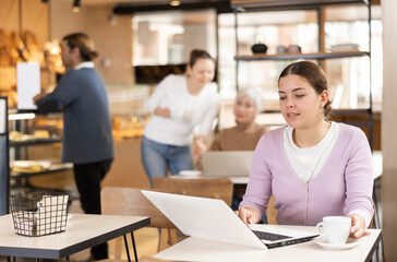 Portrait of interested young girl working on laptop in cafe, sitting at table over cup of coffee and browsing emails