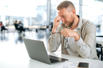 Tired exhausted caucasian man, company employee, IT specialist, sits at work desk, took off his...