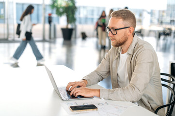 Successful motivated caucasian man with a glasses and in a casual stylish clothes, IT specialist, developer, programmer, working in a laptop on a new project in the office, developing a new program