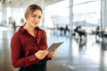 Portrait of a gorgeous successful caucasian woman, business lady, employee, broker, dressed in elegant cloth, stands in modern office, holding tablet, looking and posing at the camera, smiles friendly