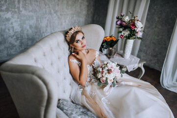A beautiful, young blonde girl bride with a diadem, a crown on her head sits in a beige sofa in a...