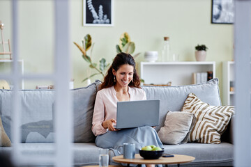 Front view of smiling young woman using laptop while sitting on sofa at home and using internet or...